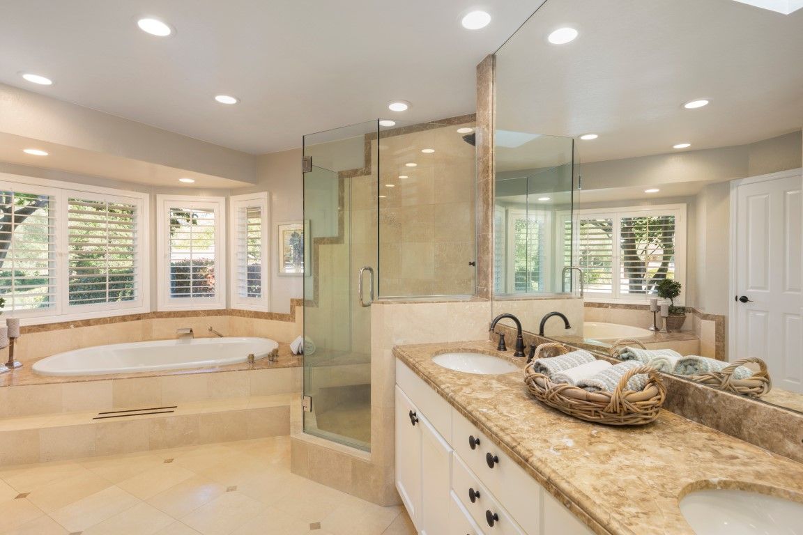 Cream marble bathroom with a corner glass shower, drop-in tub under bay windows, and a double vanity