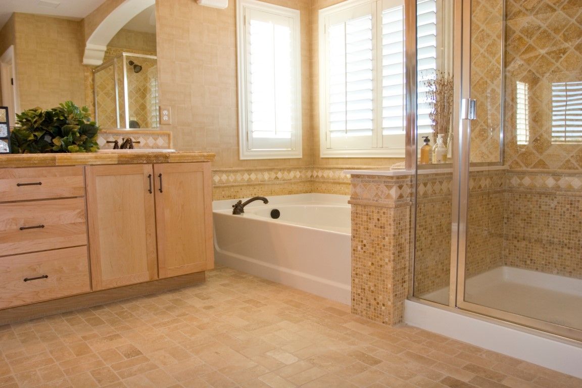 Remodeled bathroom with travertine tile floor and walls, an alcove tub, a glass shower, and a maple vanity